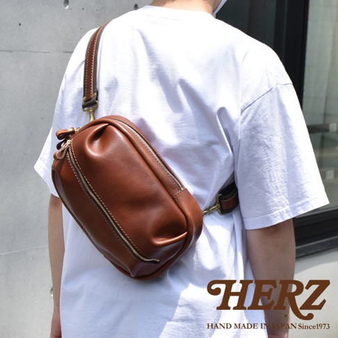 日本HERZ手作咖啡色皮革立體運動斜背包，背於身穿白色T恤的人身上，包包上印有HERZ品牌字樣。