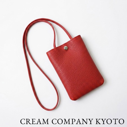 奶油色背景上呈現一款由 CREAM COMPANY KYOTO 出品的紅色皮革隨身手機包。該包款採用柔軟細緻的紋理牛皮革製成，造型簡約俐落，配有細長皮革肩背帶與單顆金屬扣環開口，底部呈現圓潤包覆感，展現日系職人手作的簡潔質感。