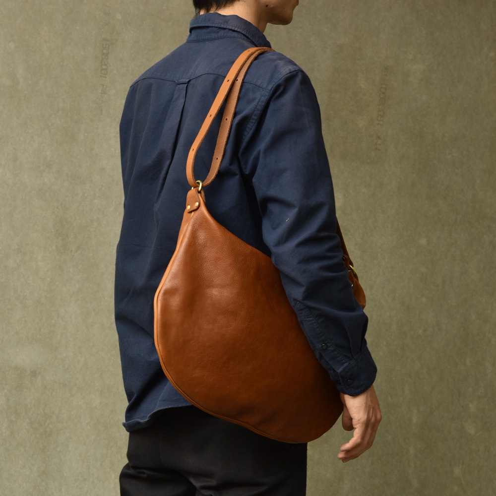 一位男性身著海軍藍襯衫，斜背一個棕褐色牛皮製的單肩半月包。包包為大尺寸，材質呈現自然的皮革紋理，肩帶為可調式設計。