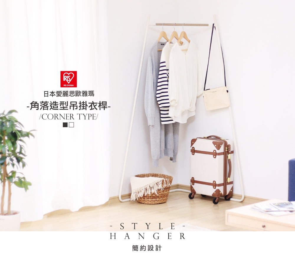 白色金屬材質的角落造型吊掛衣桿，上方掛有灰色長袖毛衣、條紋長袖上衣和白色長袖上衣，右側掛有一只米色側背包。衣桿旁有一個草編籃，籃內放有白色毯子，地面上擺放著一個米白色行李箱，箱身有棕色皮革裝飾。