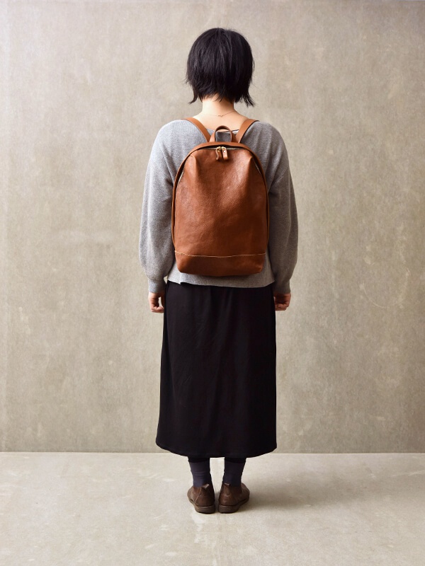 棕色皮革後背包，設有金色拉鍊和簡約褶皺設計，背在穿著灰色上衣和黑色長裙的模特兒身上。