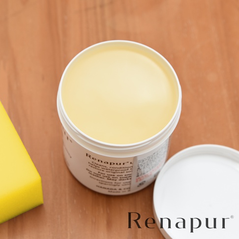 一個圓形白色罐裝的皮革保養油，內含淡黃色膏狀物。罐蓋打開，置於一旁。罐身標籤有英文說明，旁邊有一個黃色海綿。背景為木紋桌面。