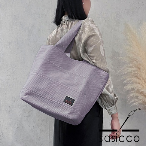 淺紫色三河木綿刺子織大容量托特包，帶有橫向條紋和黑色品牌標籤，由女性肩背。