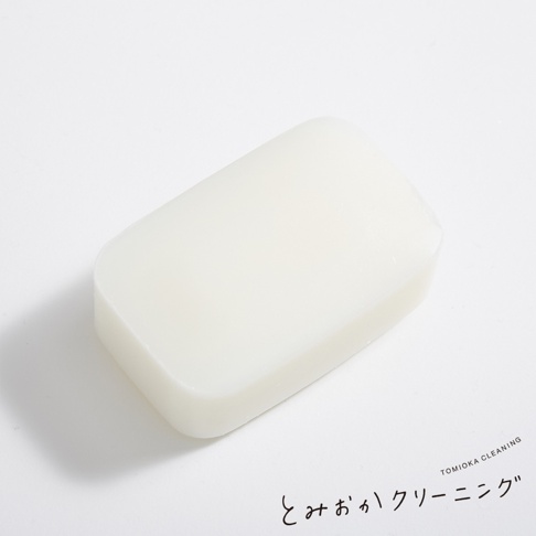 一塊白色、長方形、邊緣圓潤的洗衣皂，置於白色背景上，右下角有黑色日文手寫字體標示「とみおかクリーニング」及英文「TOMIOKA CLEANING」。