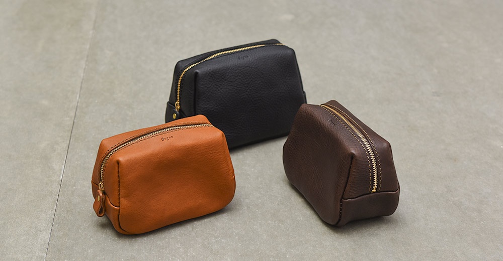 三個不同顏色的皮革拉鍊收納包放在灰色地板上，左邊為棕色，後方為黑色，右邊為深咖啡色，收納包表面刻有細小的 Organ 品牌字樣，展現出職人手作的質感。
