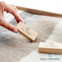 一塊淺棕色、帶有「KUSU HANDMADE ECO BLOCK」蝕刻字樣的木條，由手指拿起，置於淺色與深棕色格紋織物上。另一塊相同的木條放置於織物上。