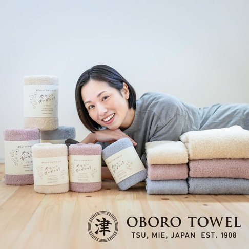 一女子側躺，身後擺放著多捲不同顏色（米白色、淡粉色、淺灰色）的有機棉毛巾，毛巾質地蓬鬆柔軟，部分毛巾帶有日文標籤。