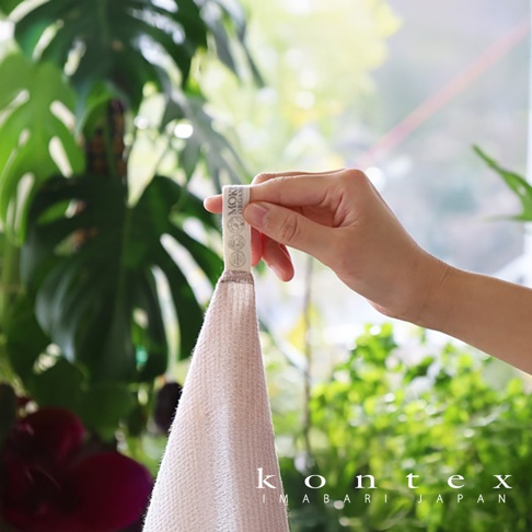 手持一條淺粉色、材質為有機棉的MOKU長毛巾，毛巾上有品牌標籤，背景為綠葉植栽。