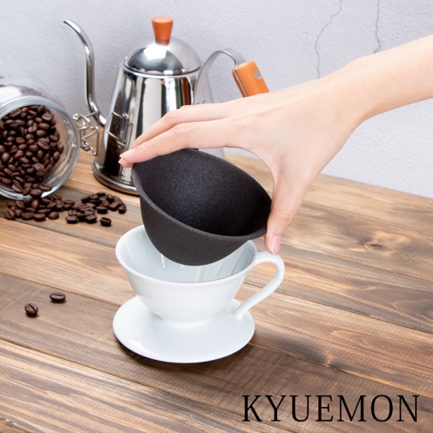 一隻手正將一個黑色、帶有細緻紋理的濾杯，放置在一個白色陶瓷濾杯組上方。背景有不鏽鋼咖啡壺和裝有咖啡豆的玻璃罐。