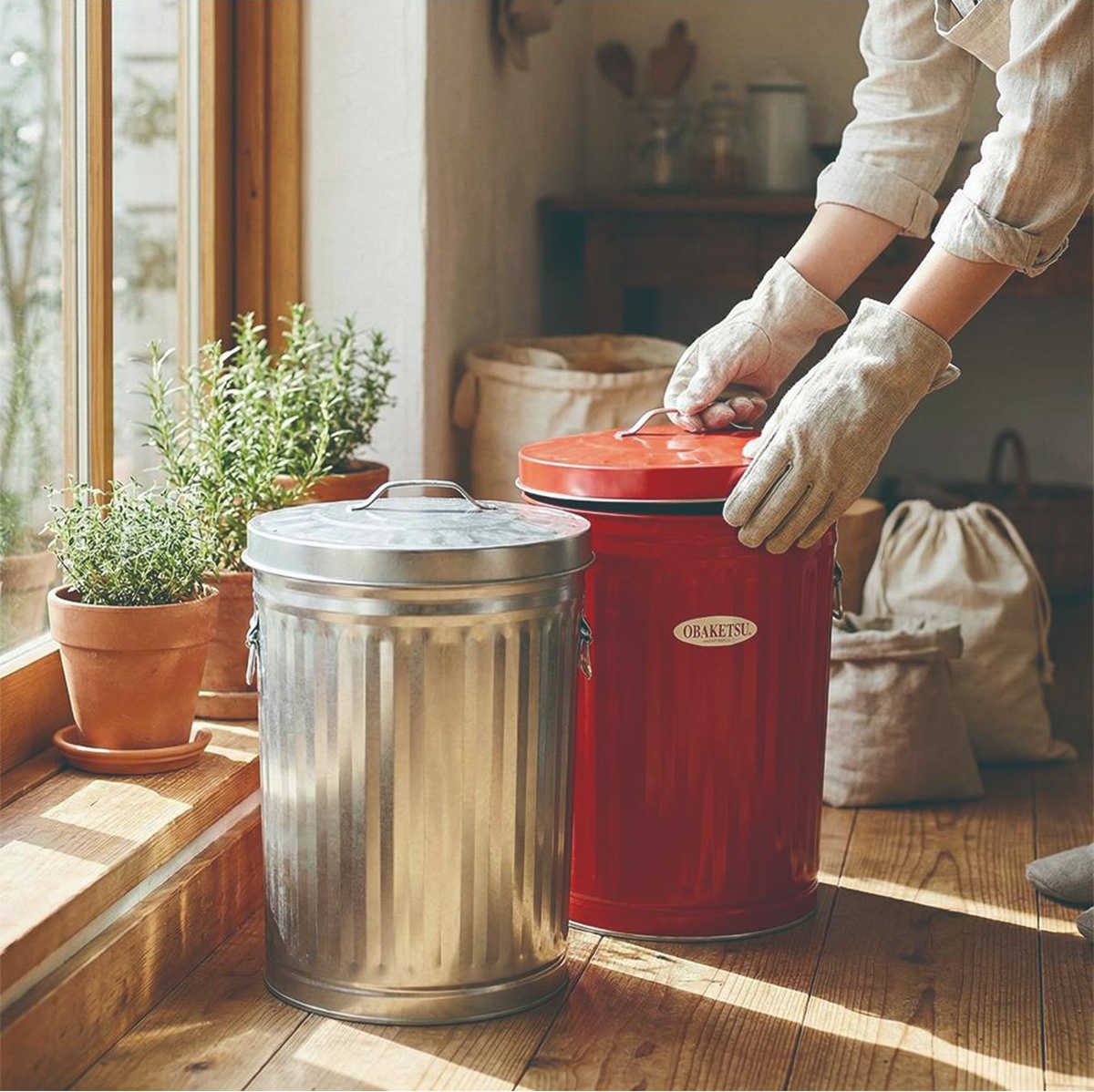 模式B： 窗邊擺放著一個銀色和一個紅色OBAKETSU手工製垃圾桶，旁邊有盆栽植物，一人戴著手套準備將紅色垃圾桶的蓋子蓋上。