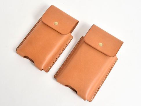 兩件淺棕色皮革手機收納包，以手工縫線和金屬按鈕閉合，設計簡潔，適合日常攜帶。