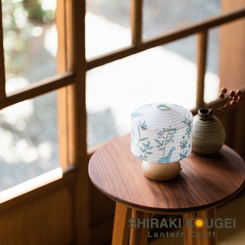 一個圓柱形白色紙質迷你提燈，燈罩印有藍色植物圖案及長頸鹿，底座為木質。