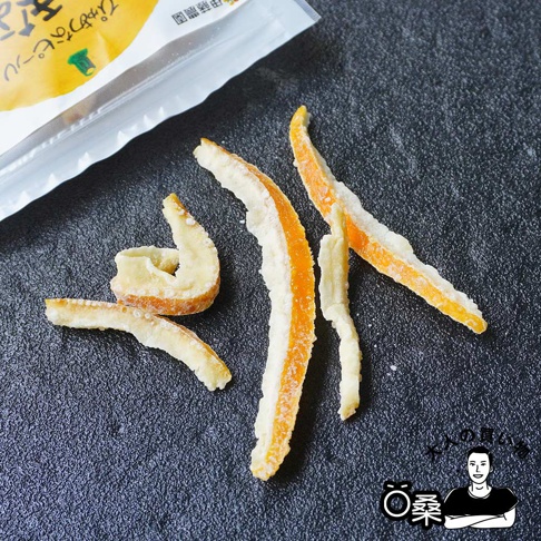 和歌山有田柑橘果皮乾，橙色果皮內層為白色，表面覆蓋一層糖霜，呈條狀或彎曲狀，擺放在深色紋理背景上。