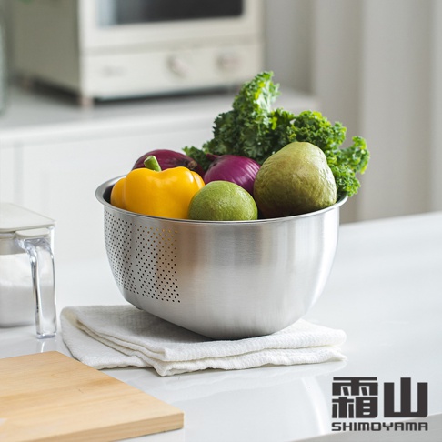 304不鏽鋼斜底瀝水料理盆，盆內裝有黃色甜椒、紫色洋蔥、綠色小球、芭樂和綠葉蔬菜，盆身佈滿圓形瀝水孔，置於白色毛巾上，背景為廚房家電。