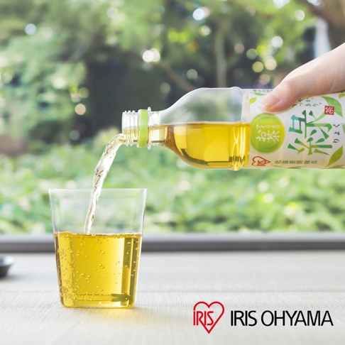 一隻手握著IRIS OHYAMA綠茶寶特瓶，將金黃色的綠茶倒入玻璃杯中，背景為明亮的戶外綠意景致，玻璃杯中呈現通透的茶色與細緻氣泡，桌面上放著印有IRIS OHYAMA品牌名稱與愛心圖示。