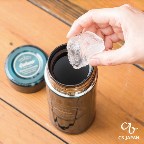 一只手正將一塊冰塊放入一個帶有「Oahu」標誌的黑色保溫杯中，保溫杯內盛有深色液體。杯蓋為藍綠色，上面印有相同的標誌。背景是木質桌面。