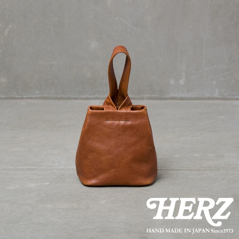 職人手作皮革Organ單柄手提包，棕色皮革，簡約設計，單提把，前方有縫線細節，置於灰色水泥背景前。