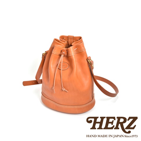 職人手作皮革束口單肩包，採用天然棕色皮革製成，附有可調節的皮革肩帶和束口設計。包身有精緻的縫線細節，正面有一個橢圓形皮革標誌。