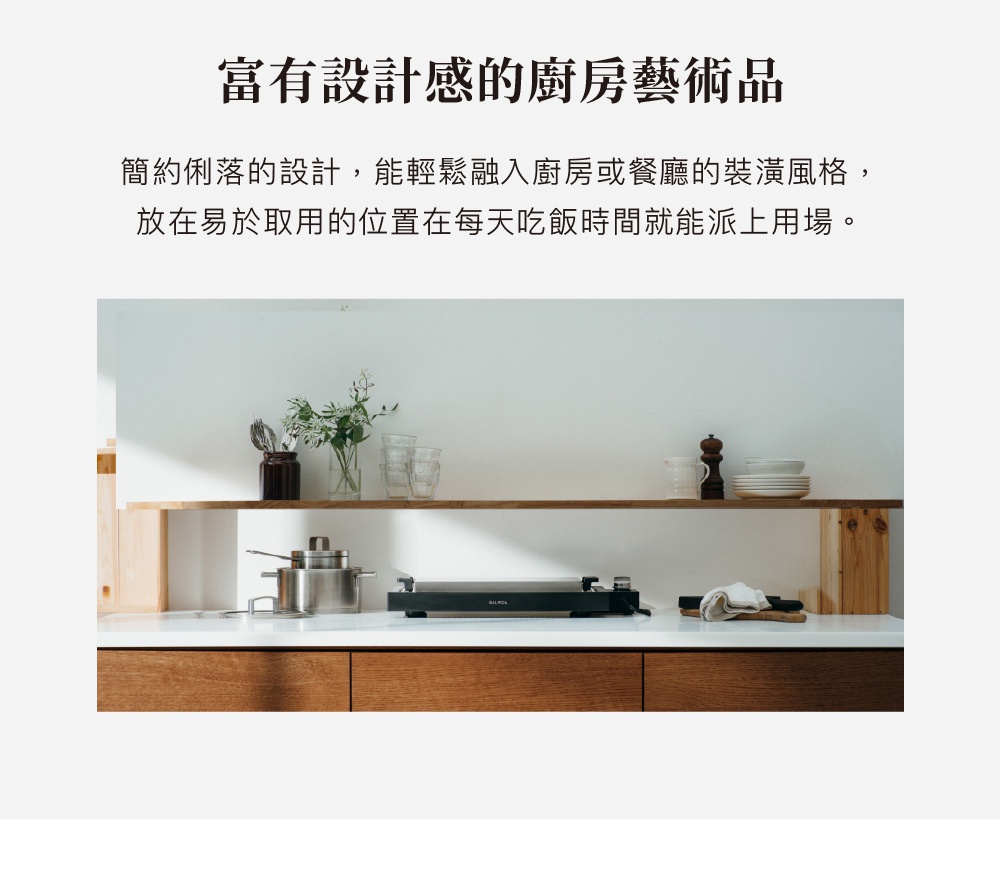 The Plate Pro 極致電烤盤 (黑色) 是一個黑色、扁平的電烤盤，放置在廚房檯面上。旁邊有不銹鋼鍋、玻璃杯、花瓶、調味瓶和疊起的盤子。