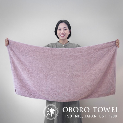 一名女性手持一條淡紫色有機棉浴巾，浴巾質地柔軟，帶有細緻的絨毛紋理。