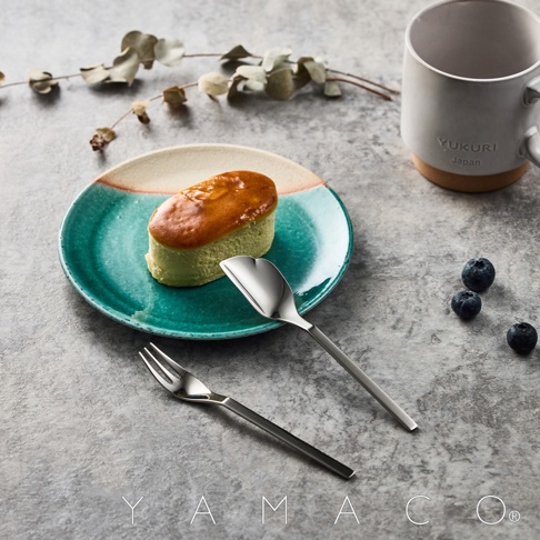 盤子上放著一個日式抹茶起司蛋糕，旁邊擺放著一套不鏽鋼蛋糕叉匙，左側有幾顆藍莓，右側有一個印有YUKURI JAPAN字樣的白色馬克杯。