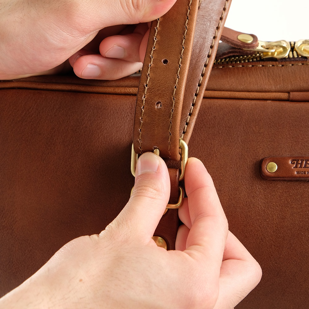 職人手作皮革肩背手提兩用公事包，展現高品質棕色皮革紋理，搭配黃銅色金屬扣環，細節處的縫線清晰可見，強調其精湛工藝。