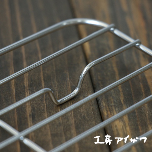 方形不鏽鋼瀝油架，由多條銀色金屬線條編織而成，呈現簡潔的網格狀結構，部分線條彎曲成U形，架設於深棕色木紋背景上。