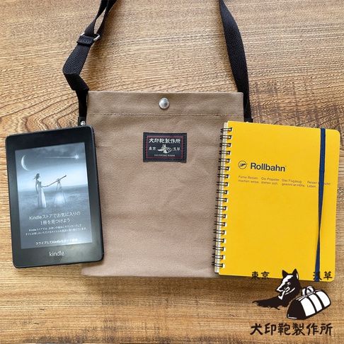 一個卡其色的帆布平板收納側背包，旁邊放著一個黃色的Rollbahn筆記本和一台Kindle電子閱讀器。
