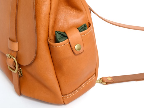 職人手作皮革屋瓦風經典雙扣後背包，以溫潤的棕褐色皮革製成，搭配黃銅色雙扣和縫線細節，側邊有一個置物小袋，內有綠色物品。
