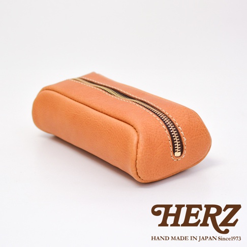 一個採用淺棕色皮革製作的HERZ品牌手工筆袋，外觀呈現圓潤的長條造型，中央配有黃銅色拉鍊，車縫線細節精緻，整體質感簡約且富有職人手作氣息，右下角印有HERZ品牌標誌。