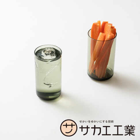 SAKAE 品牌標誌旁的日文文字「せかいをゆかにする技術 サカエ工業」，搭配左邊放著冰塊飲料的灰色玻璃杯，右邊放著胡蘿蔔條的灰色玻璃杯，整體為簡潔的產品形象照。