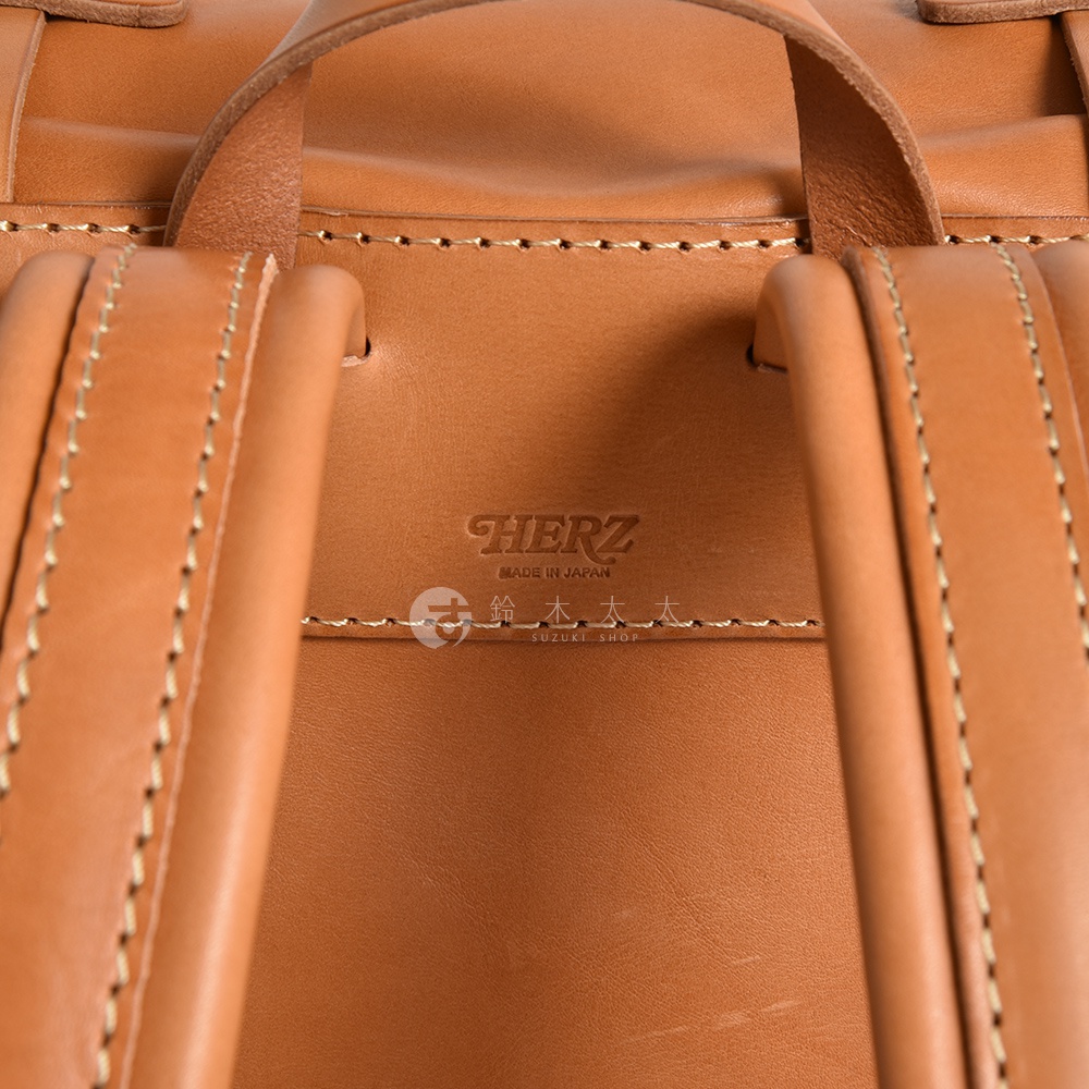 這張圖片展示了職人手作皮革露營登山用大容量後背包 R-19 的部分細節，呈現了溫潤的淺棕色皮革材質。包包的肩帶與提把部分可見品牌「HERZ」的壓印標誌，以及「MADE IN JAPAN」和「SUZUKI SHOP」等字樣，縫線精緻，質感良好。