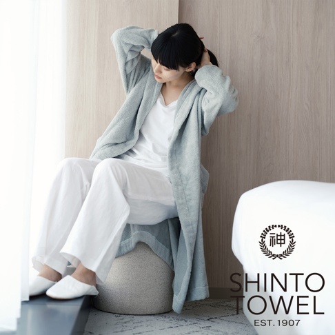 一位女性穿著淺藍色蓬鬆浴袍，內搭白色上衣與白色長褲，坐在灰白色圓形坐墊上，雙手正在整理頭髮。