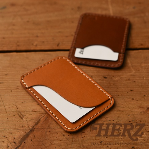 兩款職人手作皮革名片夾，一款為淺棕色，另一款為深棕色，均以白色縫線裝飾，擺放在木質背景上。