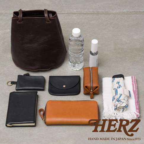 這張照片呈現日本手工品牌HERZ的皮革包款與其收納內容物。畫面主體為一個深咖啡色皮革抽繩托特包，周圍排列著錢包、皮夾、隨身小瓶、透明水瓶、折疊傘及手帕等個人日常用品，整體呈現職人手作的質感與簡約生活風格。