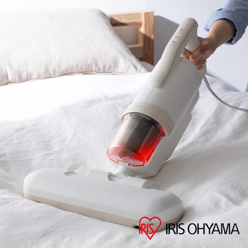 白色IRIS OHYAMA除塵蹣機，搭配透明紅色集塵筒，可見內部濾網。機身設有操作按鈕，底部連接寬扁吸頭，放置於白色床鋪上，背景為木質床頭。