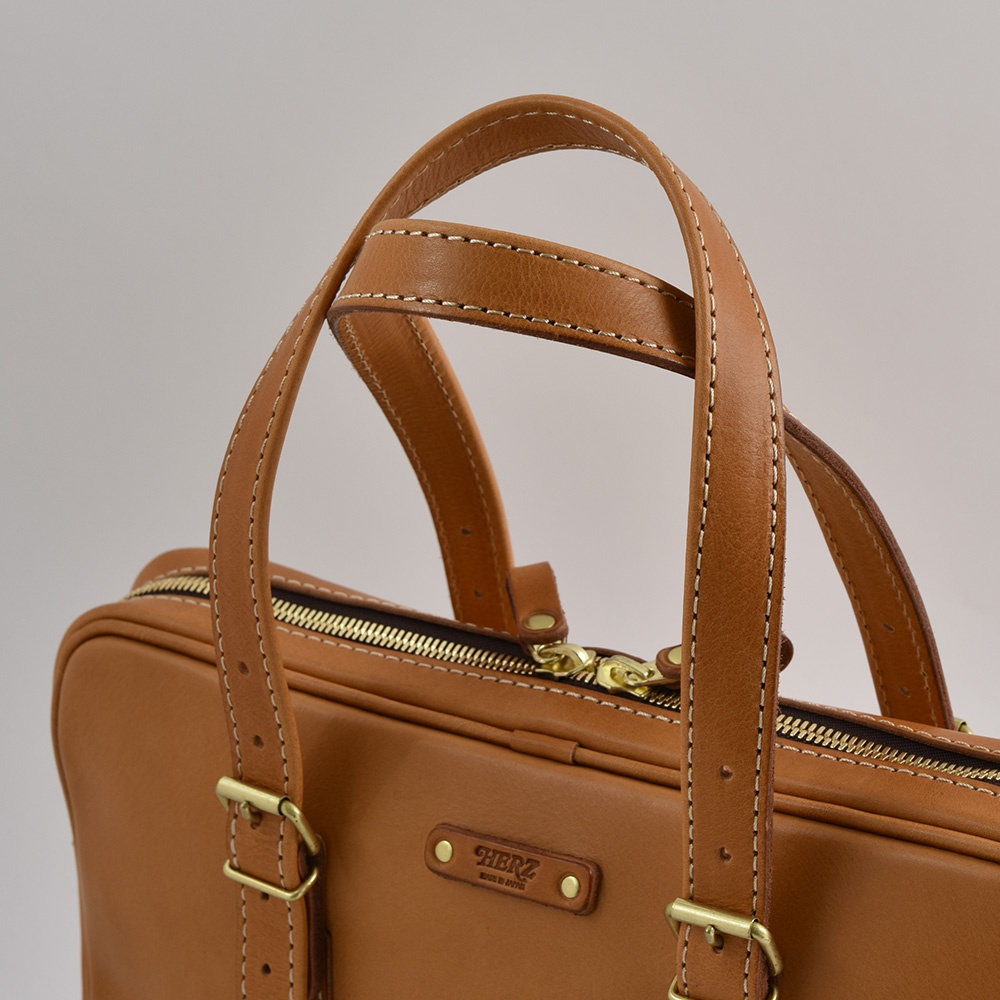 職人手作皮革公事包，採用淺棕色皮革，配有金屬拉鍊和扣環，以及手工縫製的提把與肩帶。