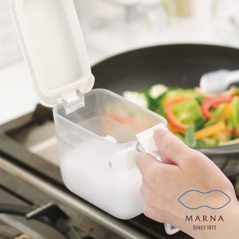 一隻手正握著 Marna 透明密封調味料盒的把手，該盒子外觀呈現半透明塑膠質感，白色上蓋處於開啟狀態，內部裝有白色粉末。背景可見瓦斯爐與正在烹飪蔬菜的炒鍋，整體呈現居家廚房的使用情境。