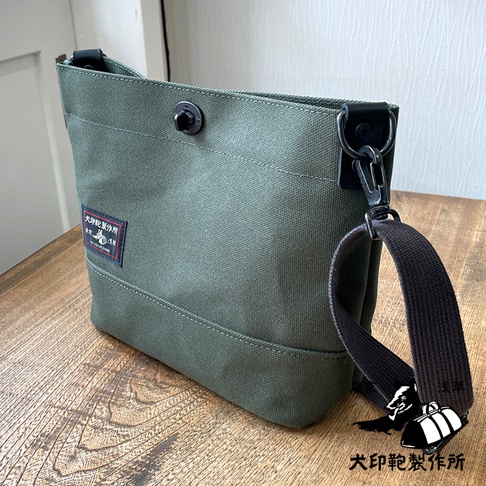 軍綠色純棉帆布迷你側背包，配有黑色織帶和金屬扣環。包身有縫線裝飾，左側印有紅色標籤，頂部有黑色按扣。