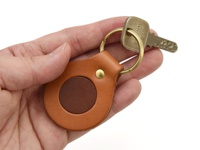 手持一只職人手作皮革圓形鑰匙圈，主體為淺棕色皮革，中央鑲嵌深棕色圓形皮革，並以金色卯釘固定，鑰匙圈處掛著一把金屬鑰匙。