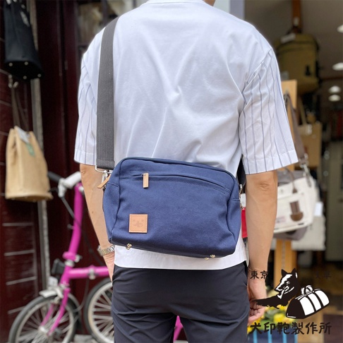側背一個藍色石洗帆布旅行側背包，包包上有一個方形的皮革標籤，此為犬印製所的商品。