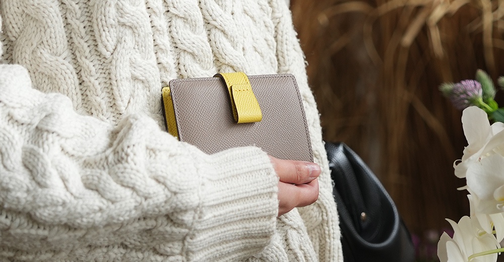 一位女士穿著厚重的白色針織衫，左手拿著一個灰色和黃色的皮革短夾，背景是模糊的乾枯植物和白色花朵。