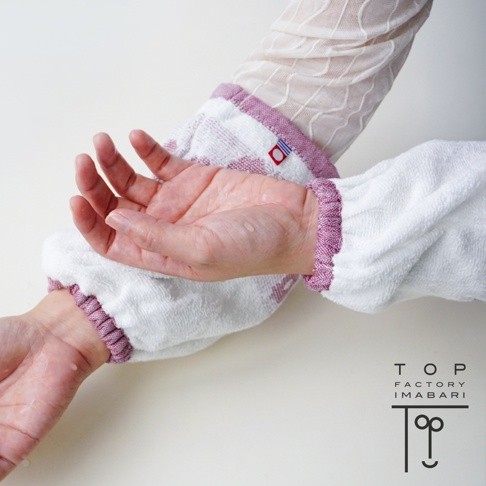 一雙穿戴在手腕上的TOP FACTORY今治純棉吸水防濕袖套，採用白色毛巾材質並配有深粉色彈性縮口，袖套上附有紅色今治認證標籤，手部與腕部帶有些許水珠，展現良好的吸水防濕功能。