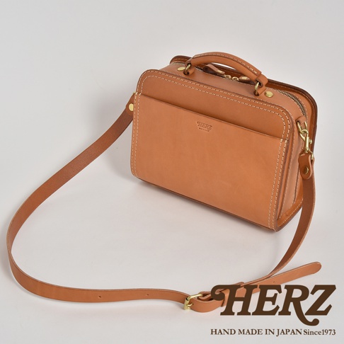 職人手作皮革兩用方形單肩包，採用淺棕色光滑皮革製成，配有頂部提把、可拆卸肩帶和拉鍊開合。包身正面有一個大口袋，上面壓印著品牌標誌。