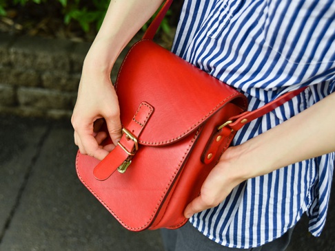 紅色職人手作皮革翻蓋迷你單肩包，配有金色金屬扣環和可調式肩帶，皮革表面有車縫線細節，由一位女性手持。