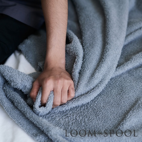 手部觸碰灰色蓬鬆柔軟的毛巾，毛巾表面有細微的絨毛感，底部印有 LOOM & SPOOL 字樣