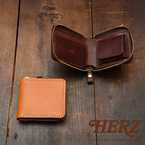 這是一張日本HERZ職人手作皮革全包式拉鍊短夾的展示圖。畫面呈現深色木質紋理桌面上，左下方放置一個封閉的淺棕色皮革短夾，右上方則展示另一個同款深褐色短夾的內部結構，展現出包含零錢袋、卡片夾層與細緻車縫線的工藝細節，右下角印有HERZ品牌標誌。