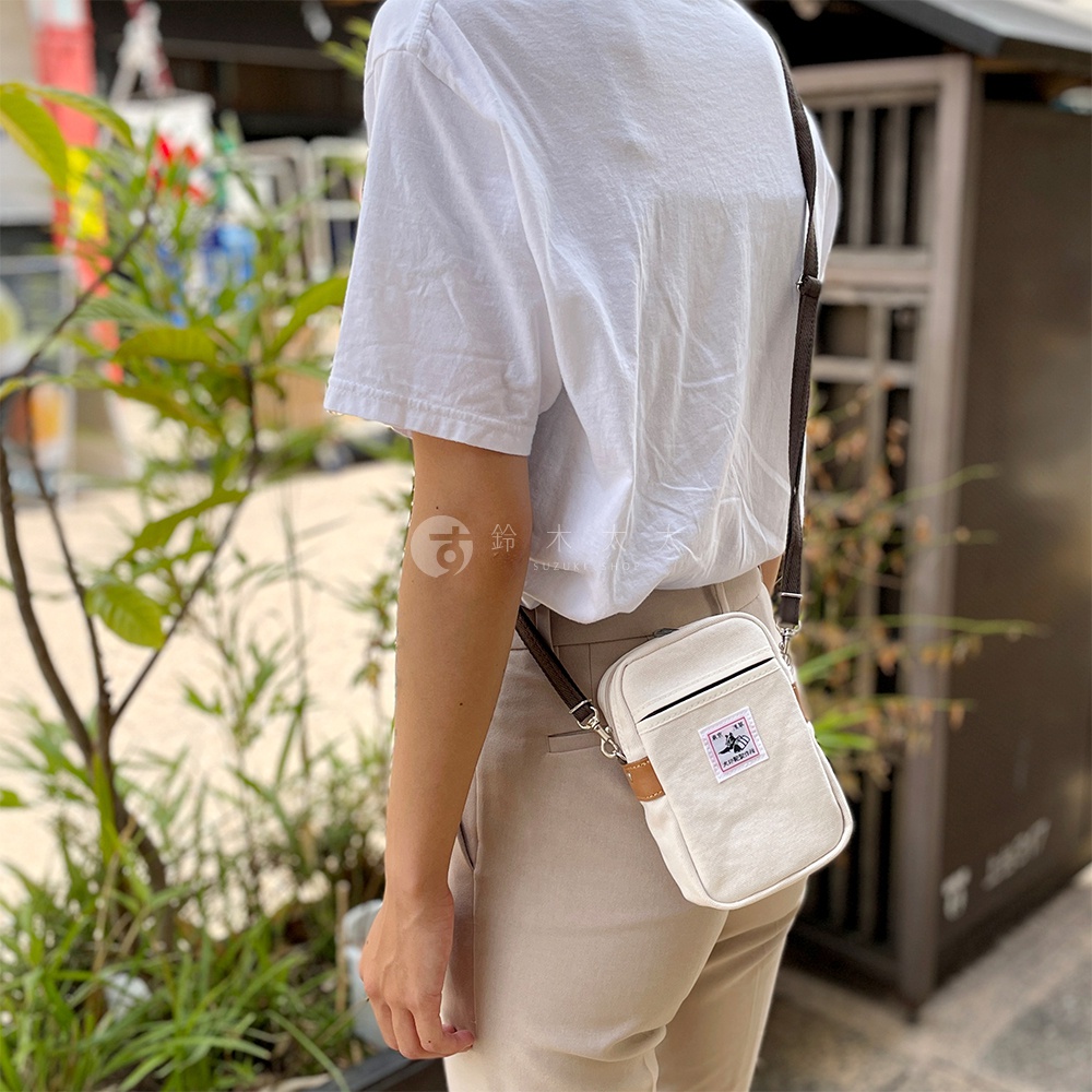 斜背在肩上的白色石洗帆布手機包，包身小巧，配有深棕色織帶和皮革飾邊，正面有一個標籤。