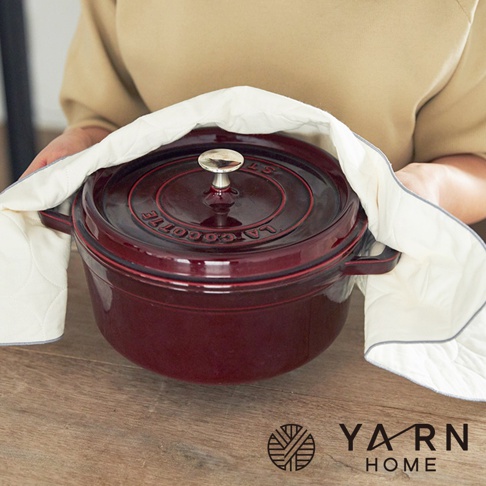 一手持著深紅色鑄鐵鍋，另一手拿著米白色絎縫紋理的棉混紗廚房拭巾，拭巾邊緣有灰色滾邊。