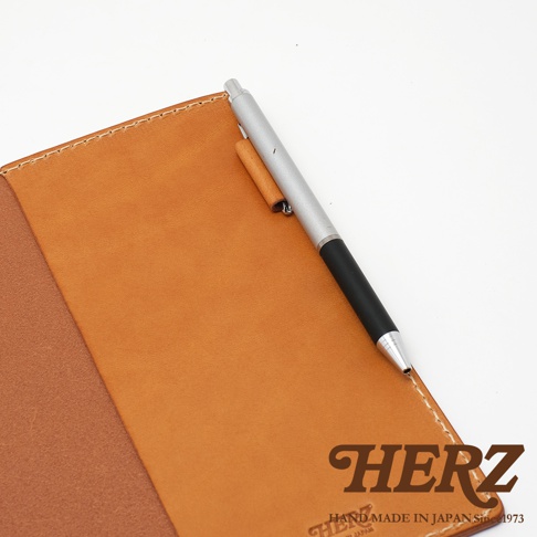 職人手作皮革B6記事本書套，採用焦糖色皮革製作，配有銀色金屬筆的筆套。